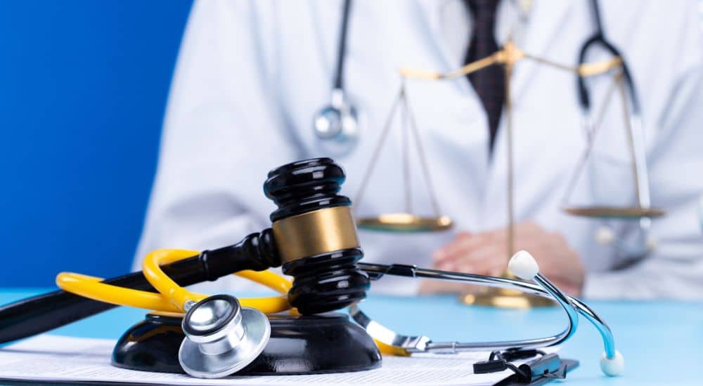 Un stéthoscope, un marteau et une balance de la justice posés sur un bureau devant un médecin en blouse blanche. Responsabilité médicale