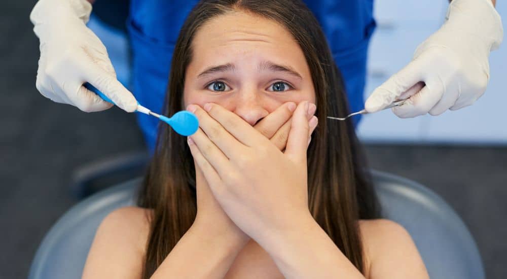 Jeune fille, dans un cabinet dentaire, les deux mains sur la bouche, mimant la peur du dentiste
